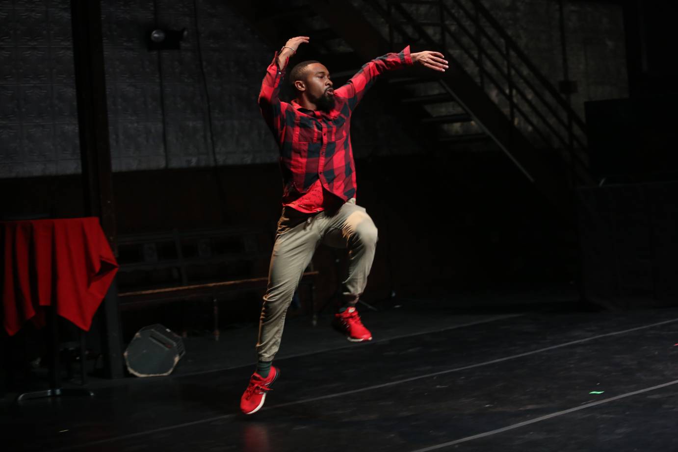 Johnie Cruise Mercer in a red and black checked lumberjack shirt, olive pants and red sneakers, jumps and reaches into the air. Behind him is a table with a red cloth and a black wooden bench